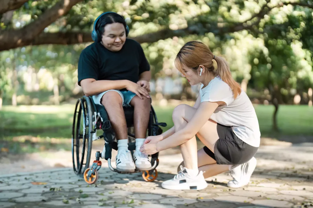 Support worker doing up shoe laces of happy disabled man in wheelchair outside at park